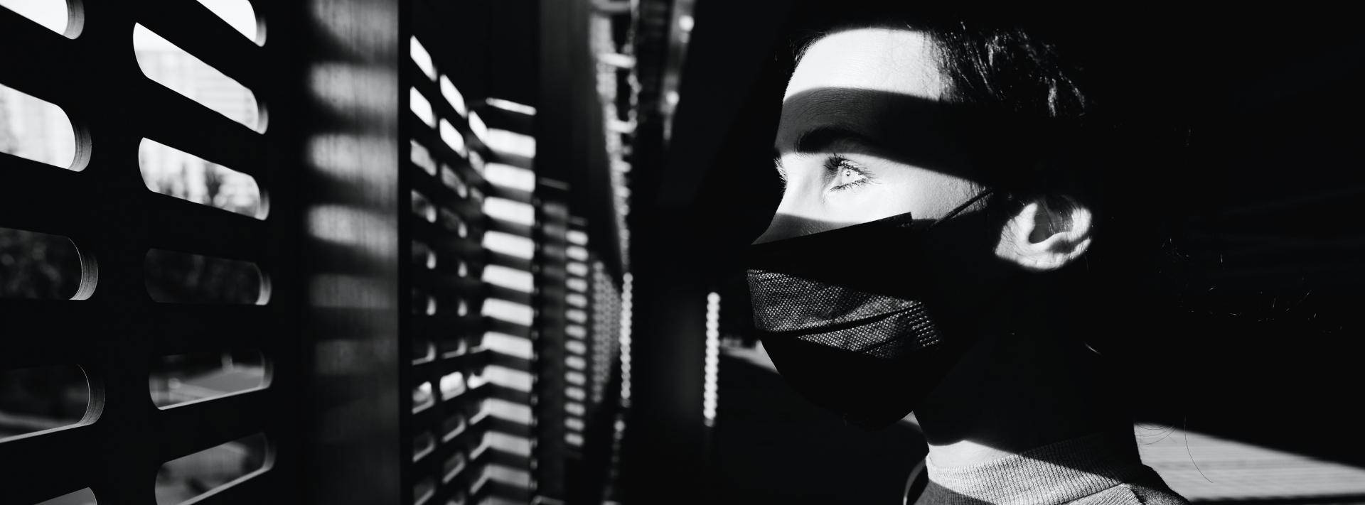 woman looking out blinds while wearing a mask - black and white image