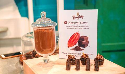 A glass vase containing new Natural Dark cocoa powder next to a visual display containing cupcakes, brownies, and a sign